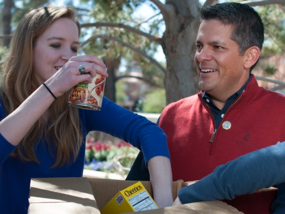 A group of people gathering nonperishable food in a cardboard box for a food drive.